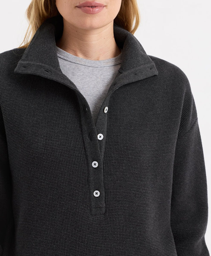 Close-up of a woman wearing a dark charcoal heather waffle-knit henley pullover with a button placket over a light gray crewneck shirt.