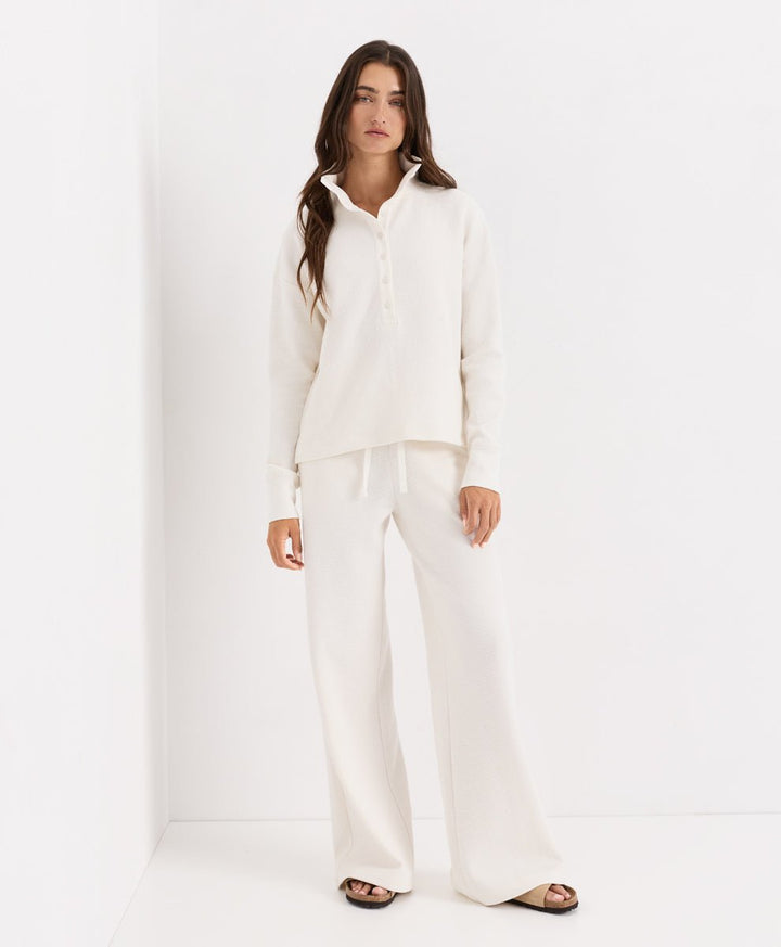 A woman modeling a matching soft ivory lounge set consisting of a long-sleeve henley and wide-leg trousers, standing against a clean white studio background.