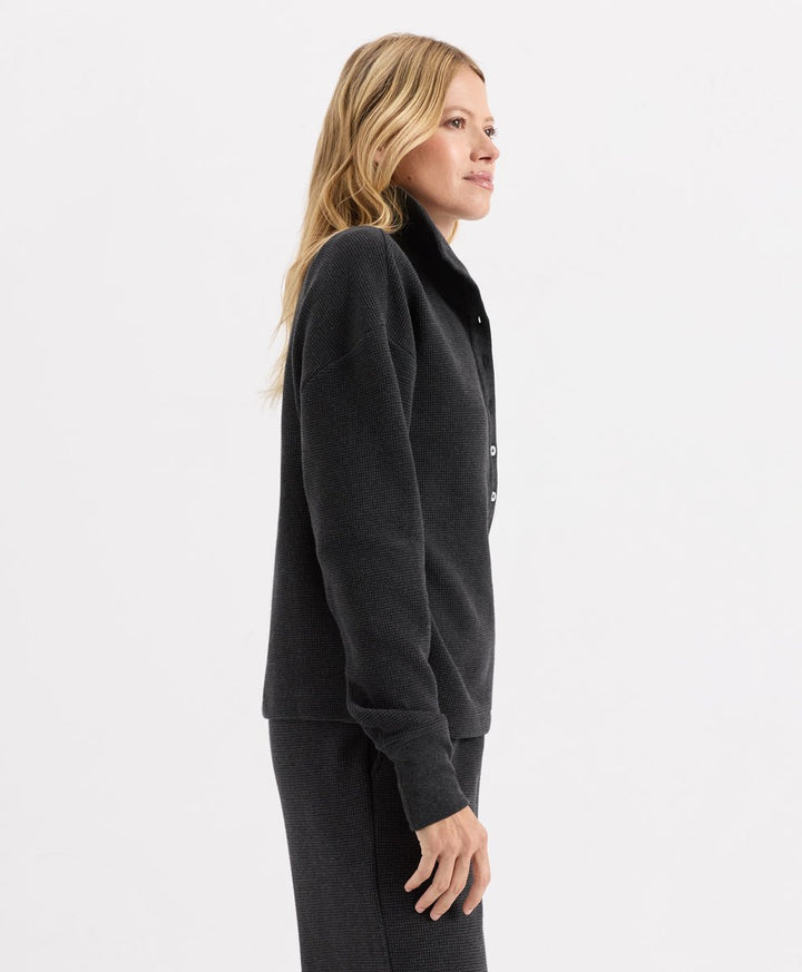 A woman shown in side profile wearing a dark charcoal heather knit button-front Henley with a collar and matching dark knit bottoms, photographed against a clean light gray studio background.