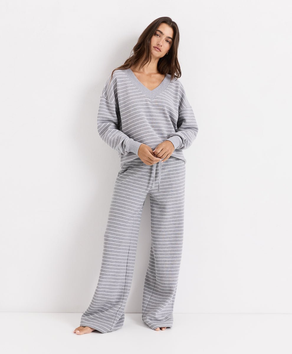 A woman modeling a matching gray and white striped lounge set, consisting of a long-sleeve V-neck top and wide-leg pants, against a plain white studio background.