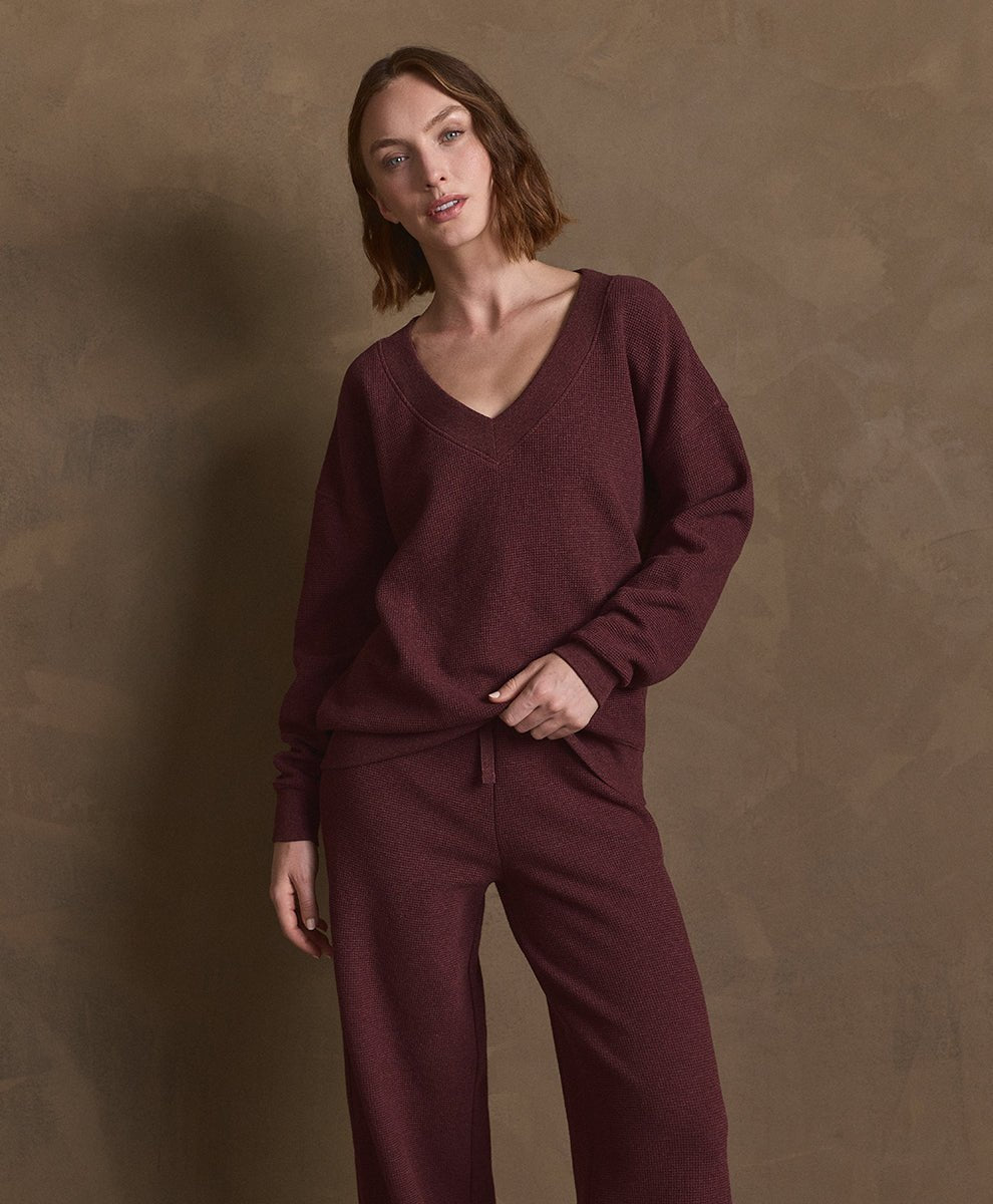 A woman modeling a matching merlot heather loungewear set consisting of a V-neck long-sleeve top and wide-leg pants, set against a textured brown studio backdrop.