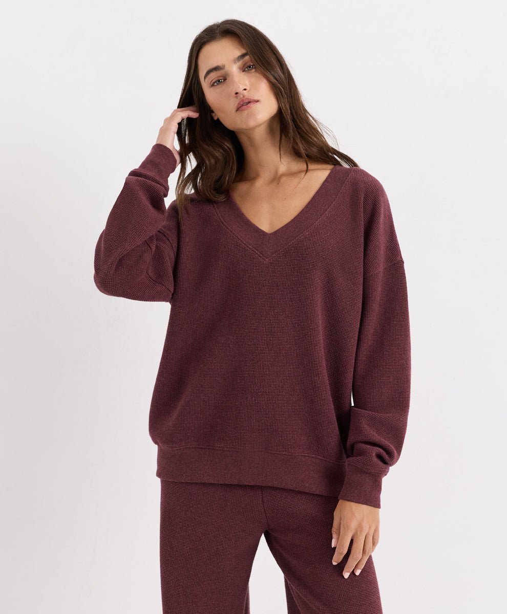 A woman modeling a matching merlot heather lounge set consisting of a V-neck long-sleeve top and coordinating pants, shot against a plain light gray studio background.