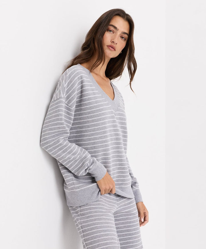 A woman wearing a light gray and white striped long-sleeve V-neck top and matching striped pants, posing against a plain white wall.