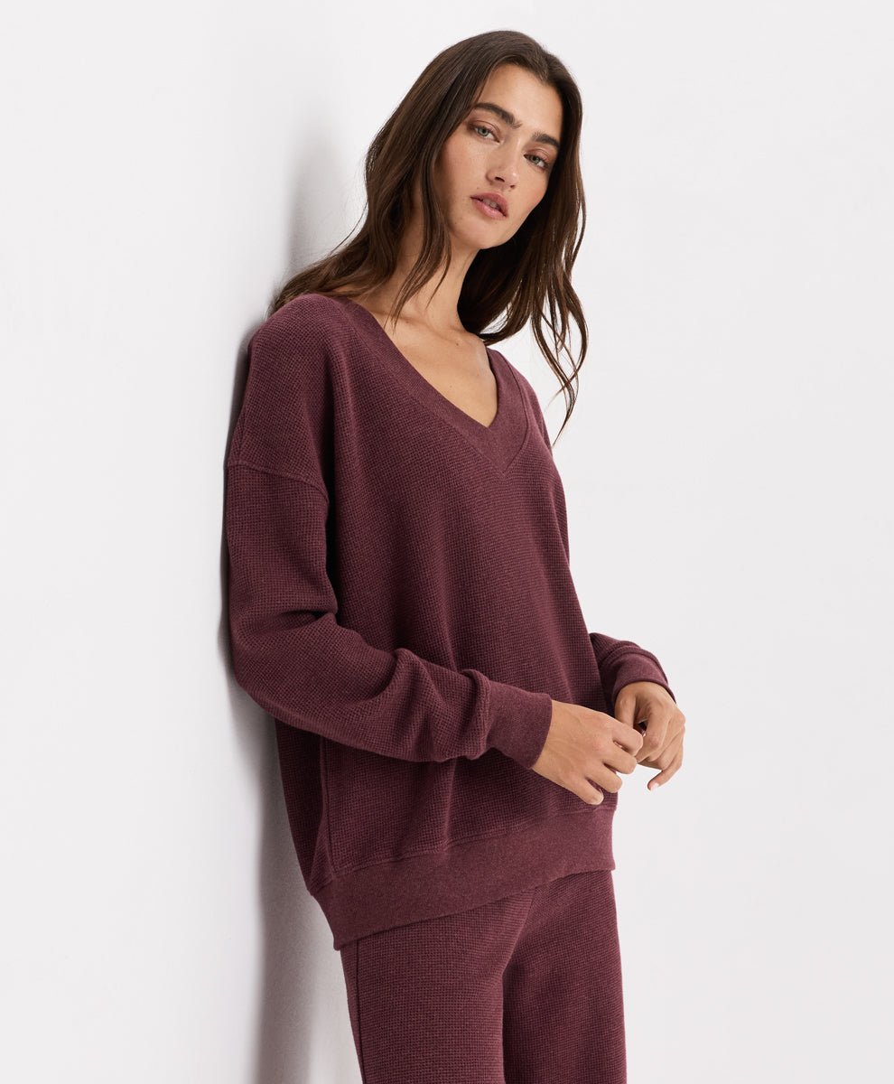 A woman wearing a matching merlot heather waffle lounge set, featuring a long-sleeve V-neck sweater and matching pants, posing against a plain white wall.