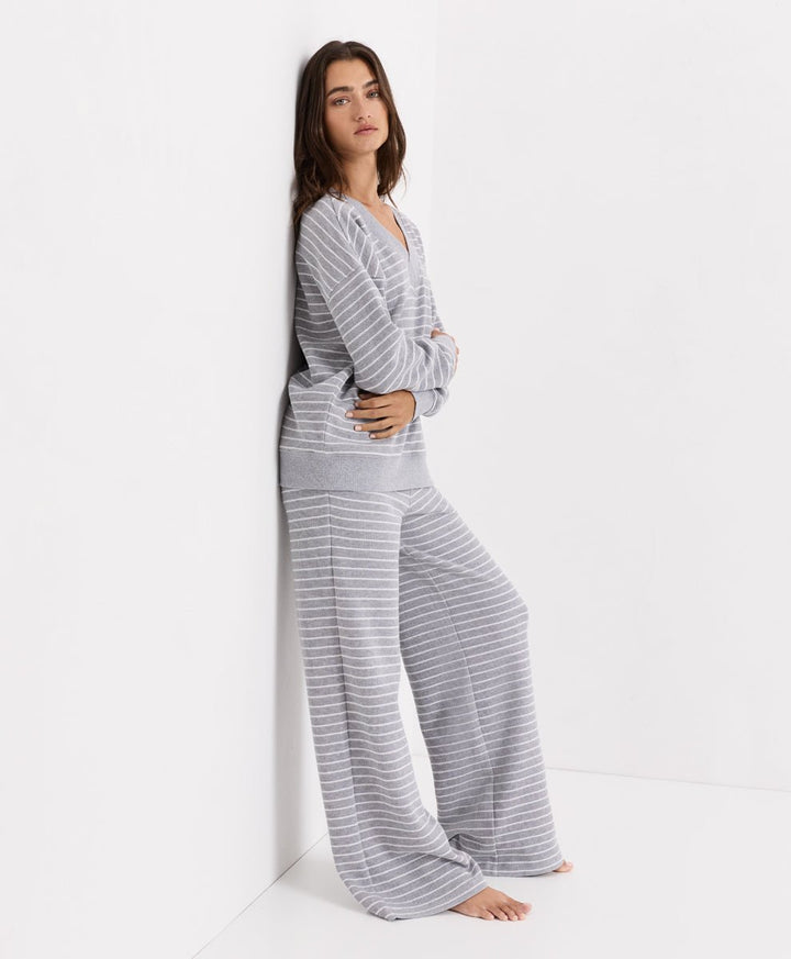 A woman wearing a matching gray and white striped lounge set consisting of a long-sleeve V-neck top and wide-leg pants, posing against a white wall in a minimalist studio setting.