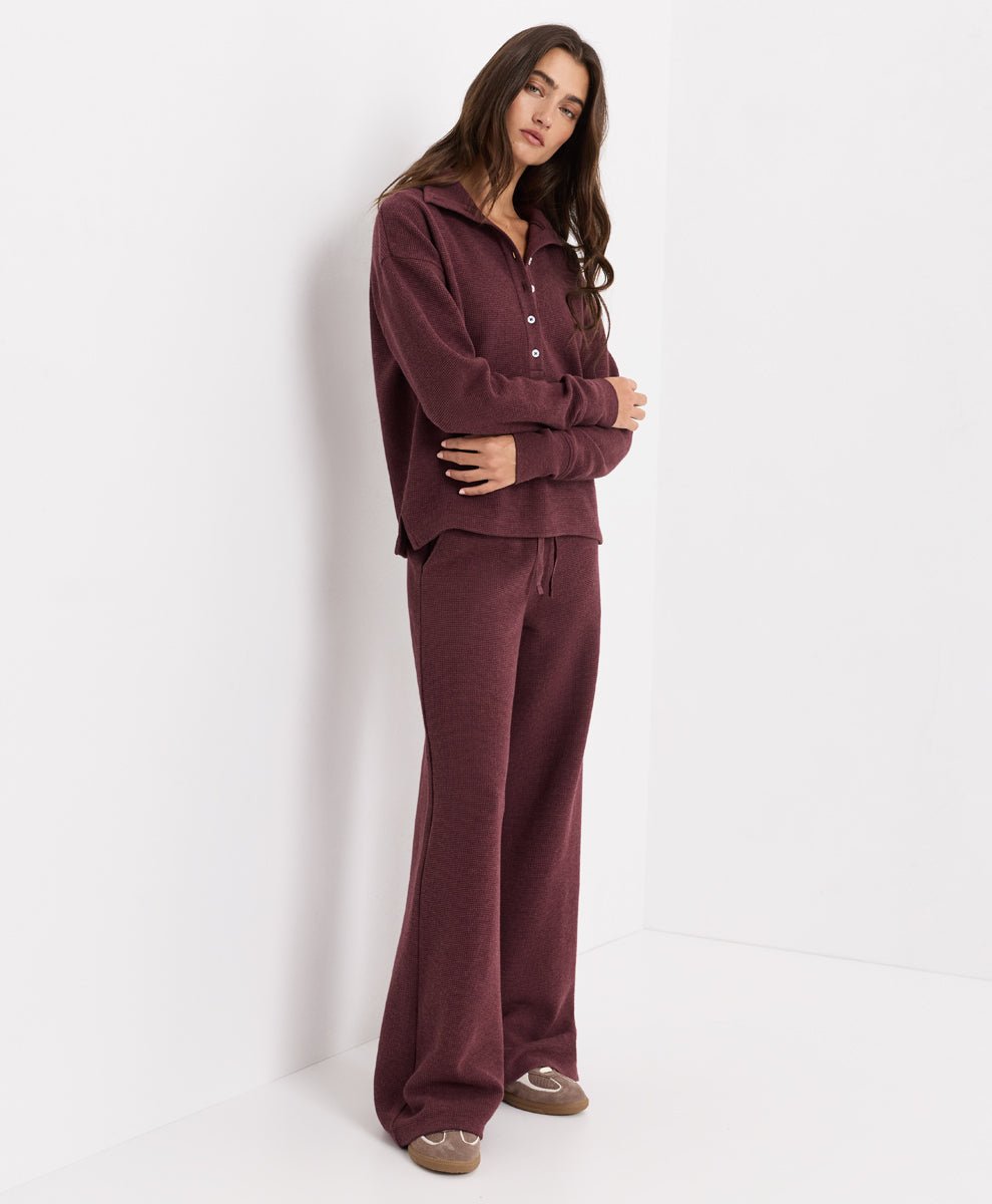 A woman modeling a matching merlot heather lounge set consisting of a long-sleeve button-front top and wide-leg pants, standing against a clean white studio background.