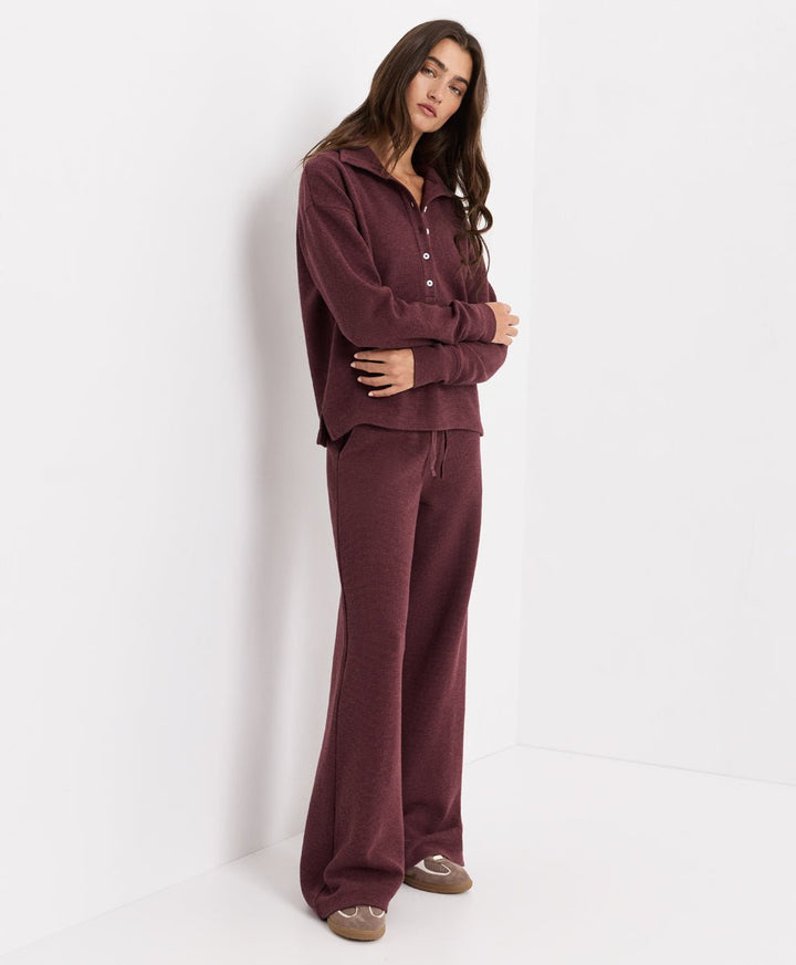 A woman modeling a matching merlot heather lounge set consisting of a long-sleeve button-front top and wide-leg pants, standing against a clean white studio background.