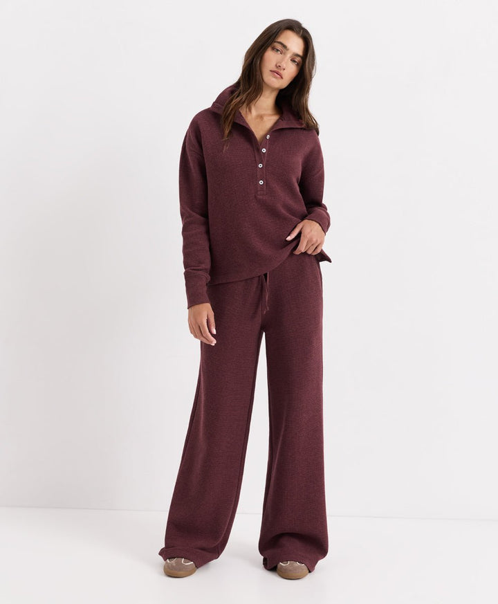 A woman modeling a matching merlot heather lounge set consisting of a long-sleeve henley top with a button placket and wide-leg pants, photographed against a clean white studio background.