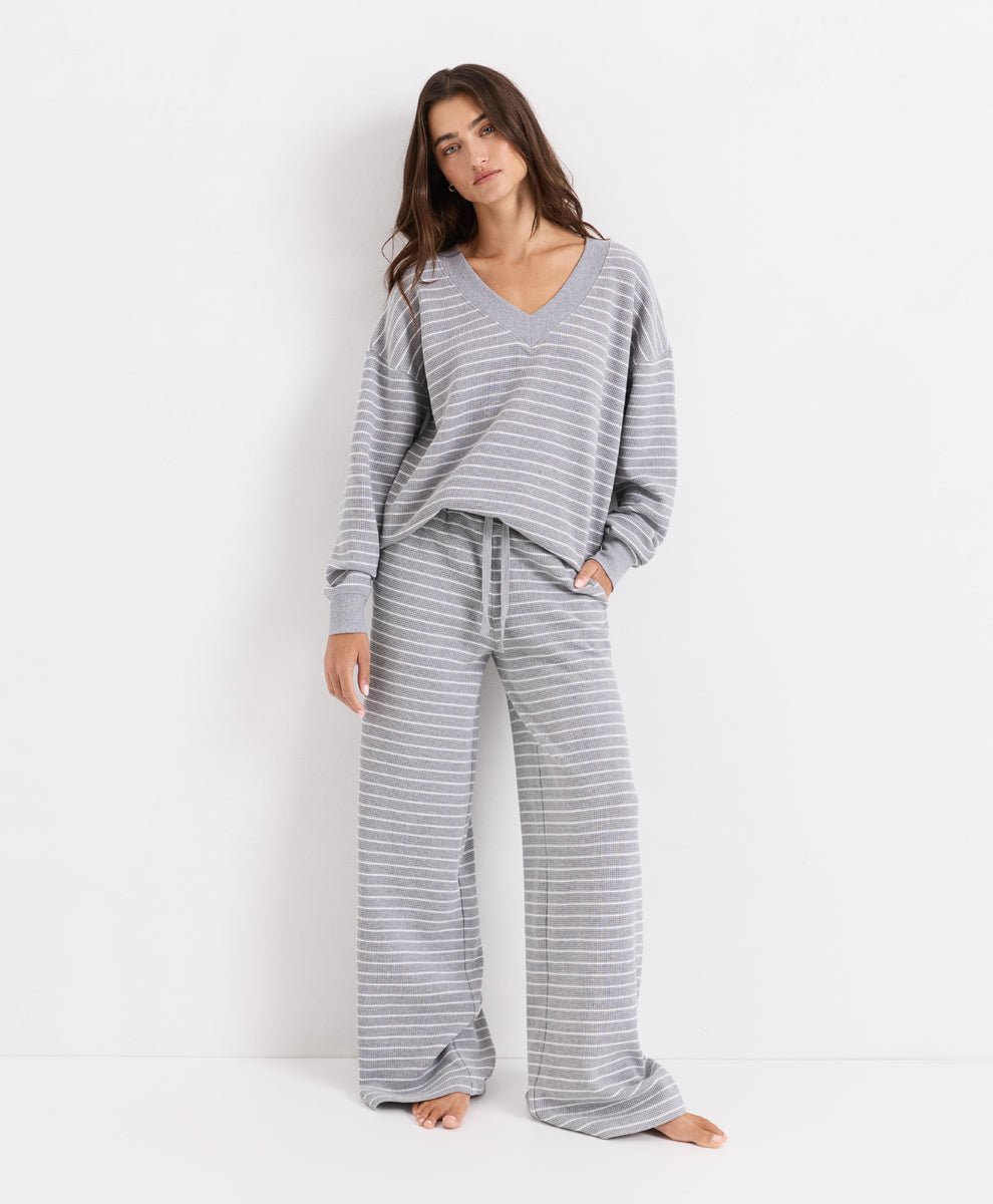 A woman modeling a matching gray and white striped lounge set, consisting of a long-sleeve V-neck top and wide-leg pants, against a plain white studio background.