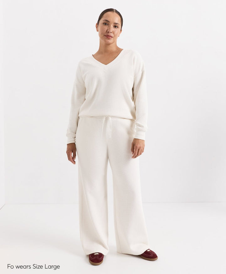 A woman modeling a matching soft ivory knit loungewear set consisting of a long-sleeve V-neck top and wide-leg pants, paired with burgundy slip-on shoes. The text &