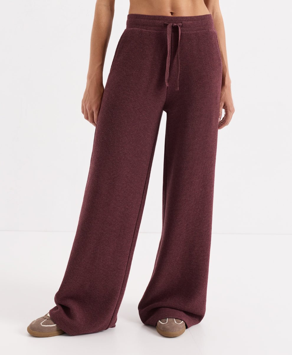 A model wearing a pair of wide-leg lounge pants in merlot heather, paired with brown slippers, against a plain white studio background.