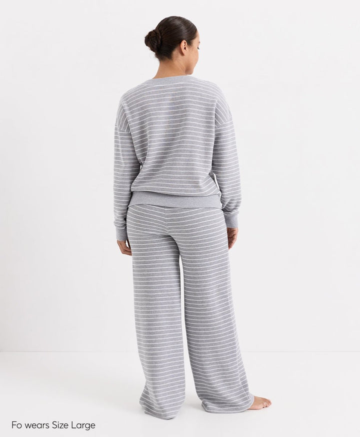 A woman stands with her back to the camera wearing a light gray and white striped long-sleeve top and matching wide-leg pants. The text &