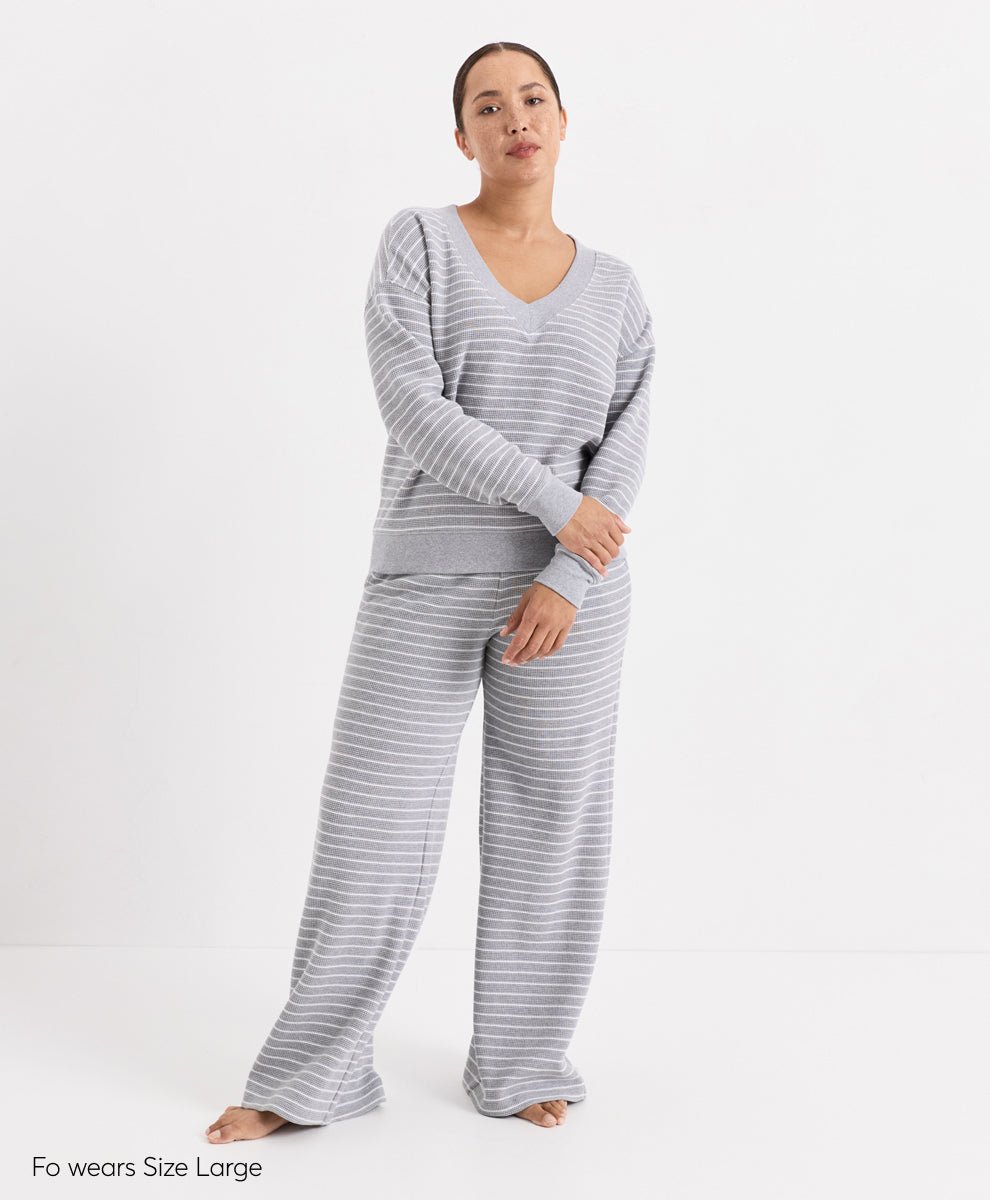 A woman modeling a two-piece loungewear set consisting of a long-sleeve V-neck top and matching wide-leg pants, both in a light gray and white stripe pattern. She is standing barefoot against a plain white studio background.