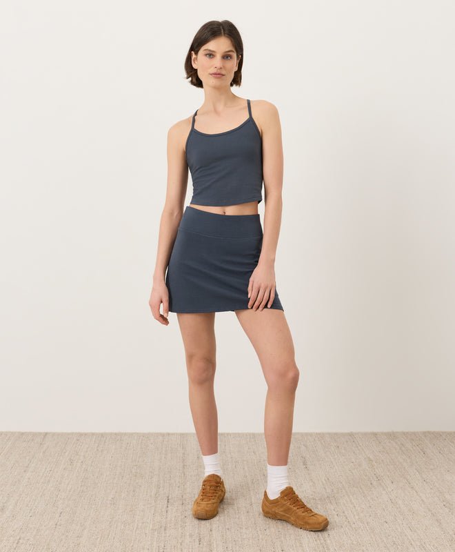 Woman wearing a steel blue tank top and a steel blue Pact skort and brown shoes on a plain background