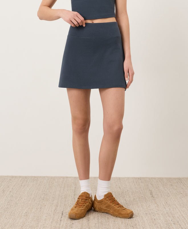 Person wearing a steel blue Pact skort and brown shoes on a plain background
