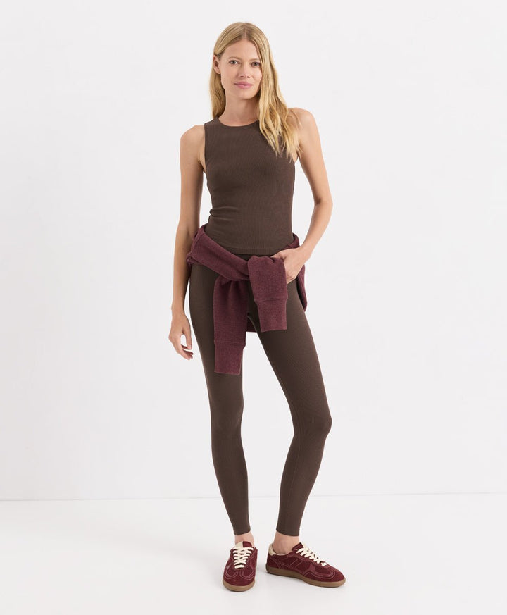 A woman modeling a sleeveless cocoa ribbed tank top, matching leggings, and burgundy sneakers, with a burgundy knit sweater tied around her waist, against a plain white studio background.