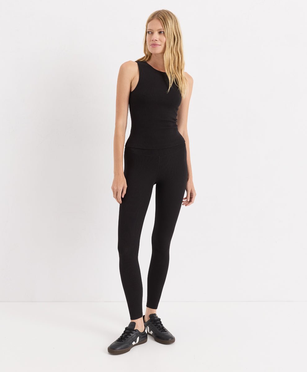 A blonde model stands against a plain white studio background wearing a black sleeveless ribbed top, black leggings, and black sneakers.