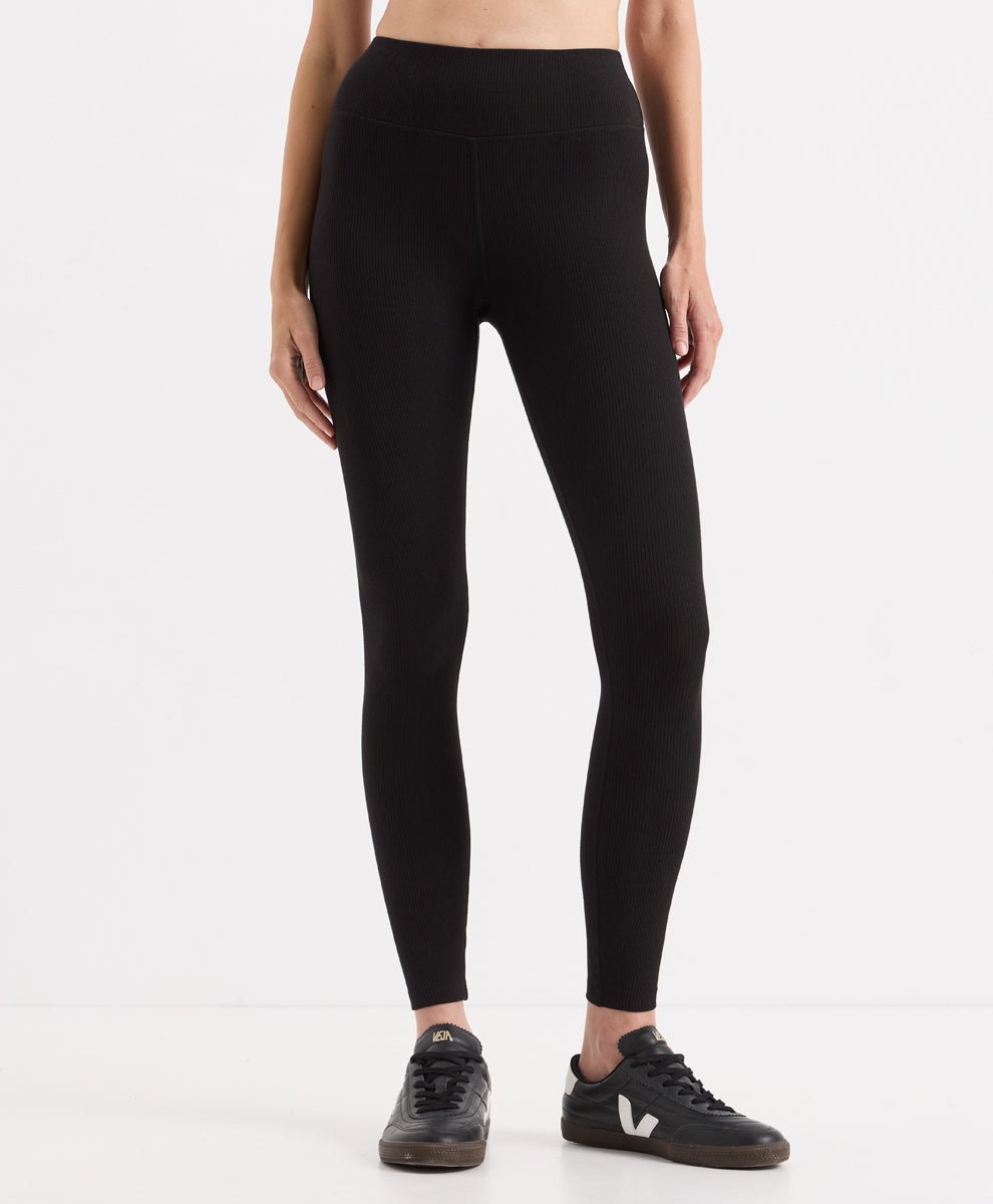 A model from the waist down wearing black high-waisted ribbed leggings and black sneakers, photographed against a clean white studio background.