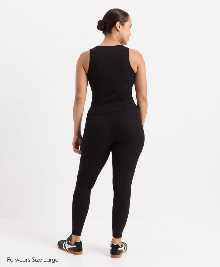 A woman shown from the back wearing a black sleeveless top and black leggings, standing against a plain white studio background. Small text in the lower left reads &