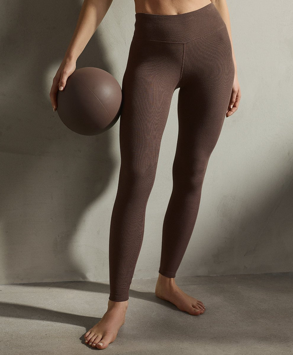 A model wearing ribbed cocoa high-waisted leggings, holding a matching brown exercise ball against a textured concrete wall.
