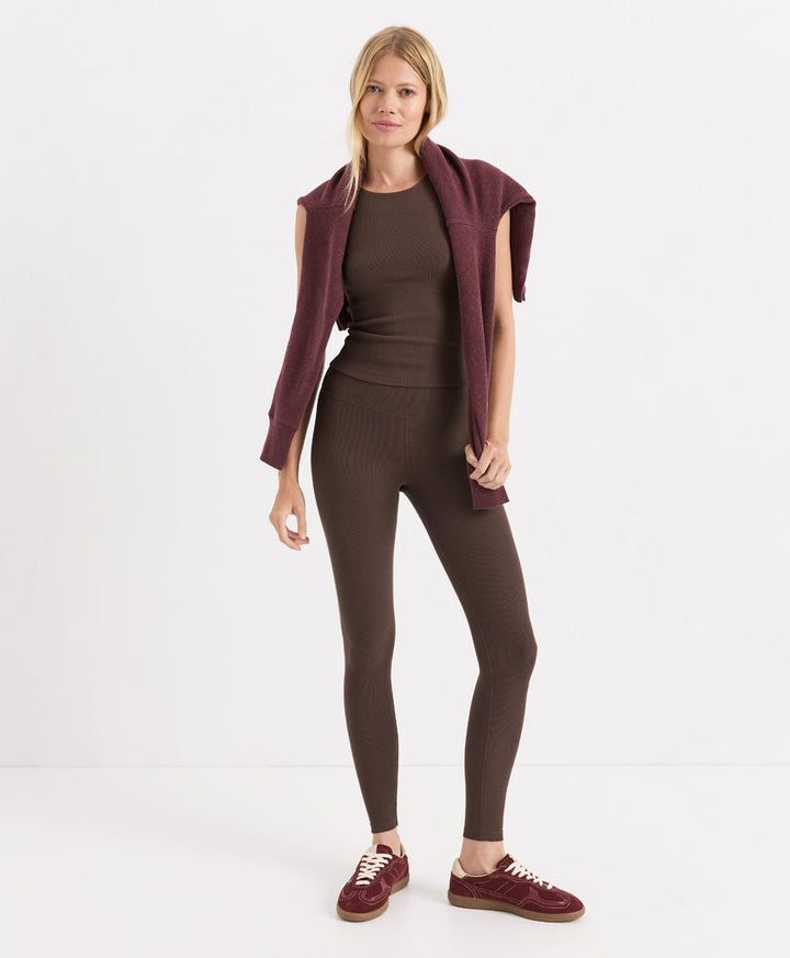 A woman modeling a matching ribbed knit lounge set in cocoa, featuring a sleeveless top, ribbed leggings, and burgundy slip-on shoes, against a plain white studio background.
