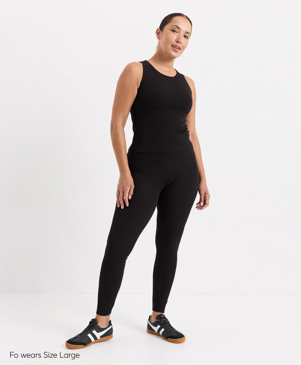 A woman modeling a black sleeveless top and black leggings set against a plain white studio background. She is wearing black and white sneakers with brown soles.