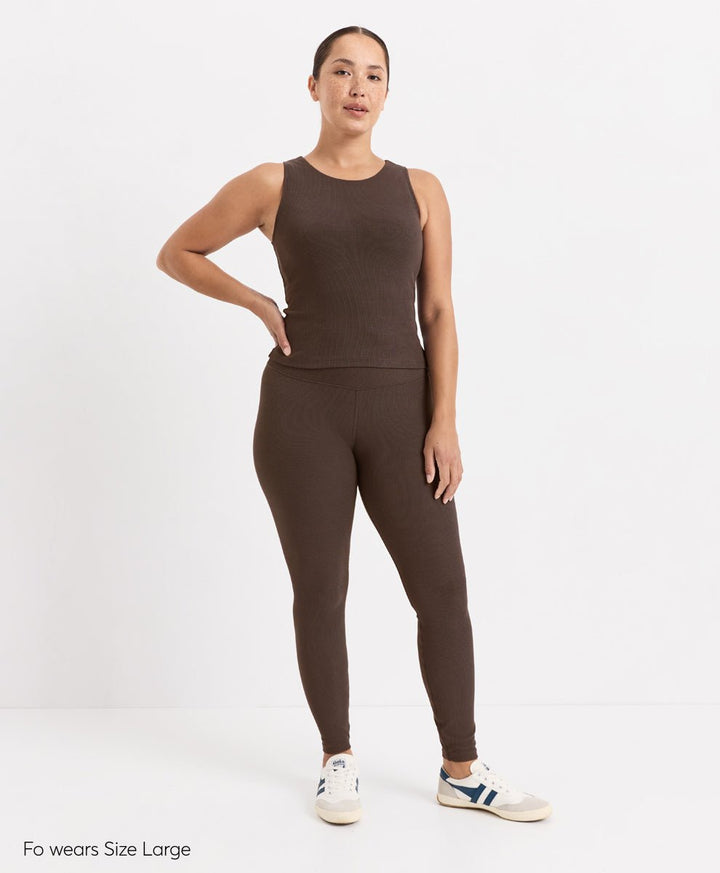 A woman modeling a matching ribbed activewear set in cocoa, featuring a sleeveless tank top and high-waisted leggings, paired with white sneakers. The text &