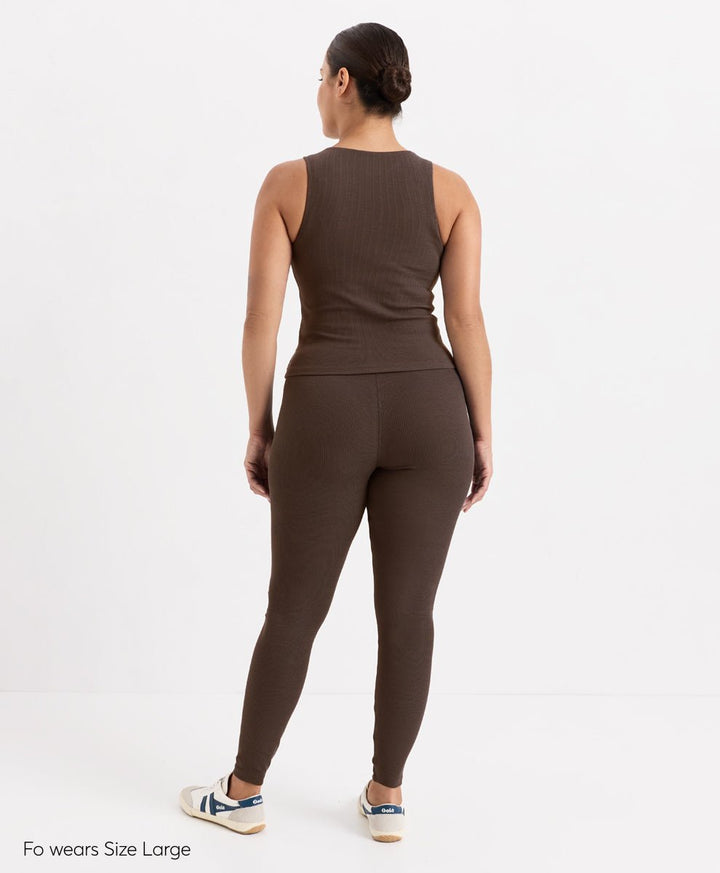 A woman shown from the back wearing a matching ribbed two-piece activewear set in cocoa, consisting of a sleeveless top and high-waisted leggings. She is standing against a clean white studio background with text in the bottom-left corner reading &