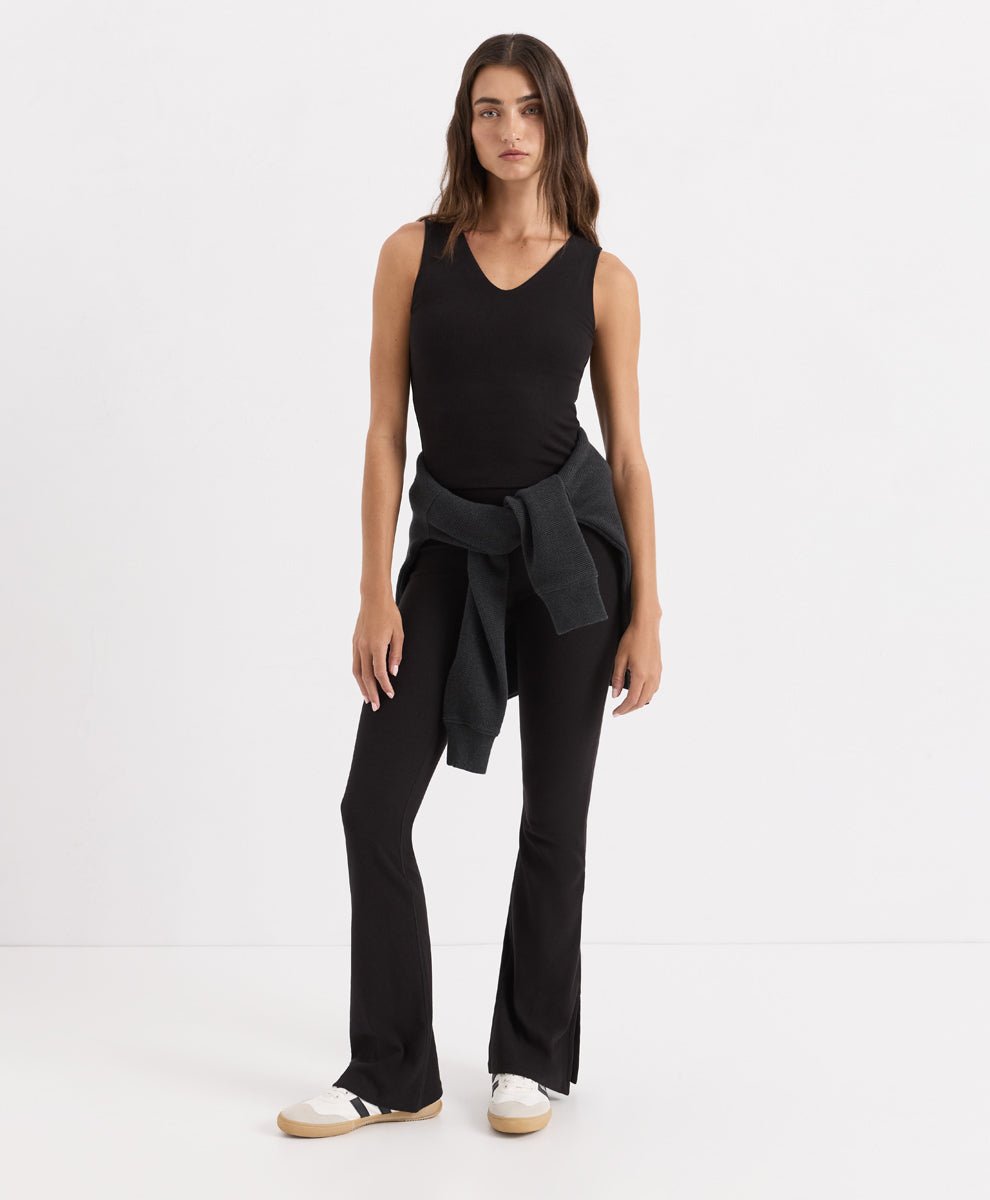 A woman modeling a black sleeveless V-neck top, black flared pants, and white slip-on shoes against a plain white studio background.