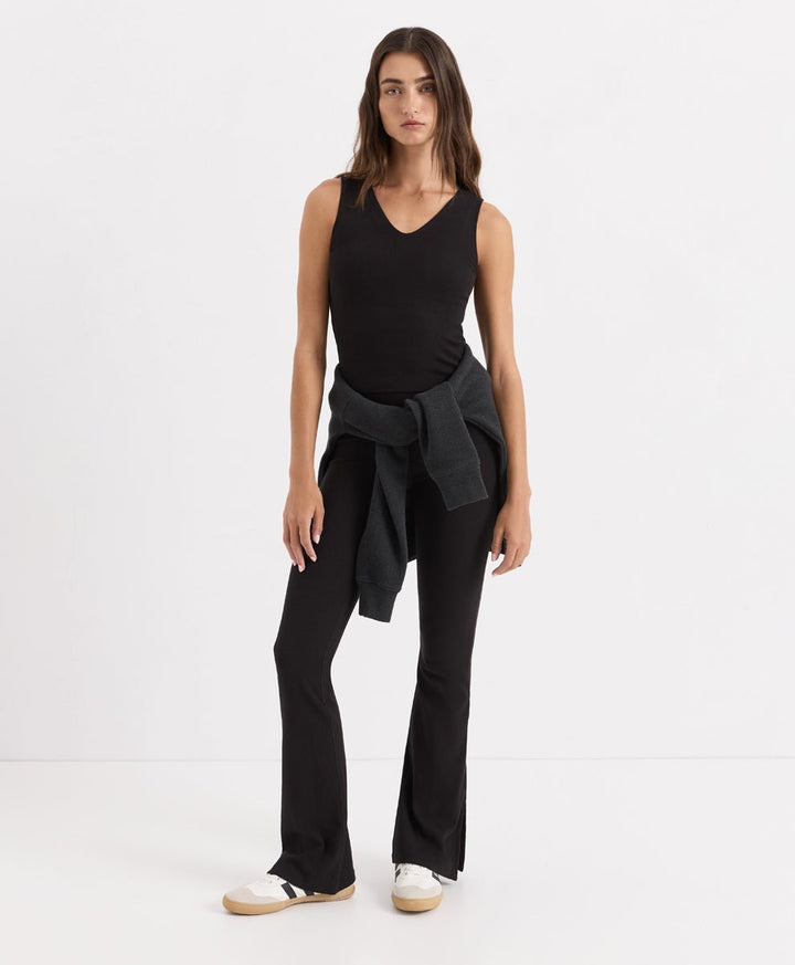 A woman modeling a black sleeveless V-neck top, black flared pants, and white slip-on shoes against a plain white studio background.
