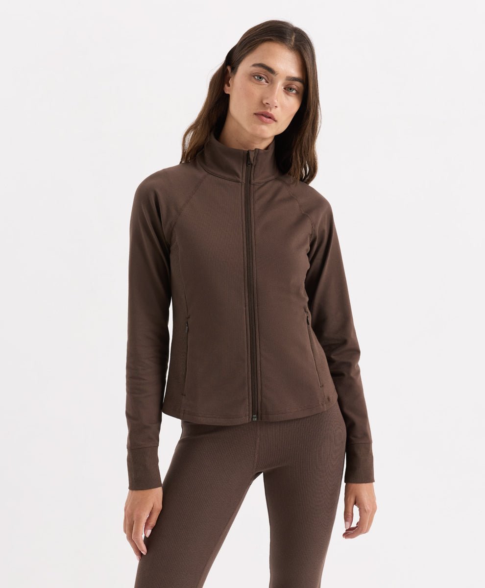 A woman modeling Pact's cocoa zip-up athletic jacket with matching brown leggings against a plain white studio background.