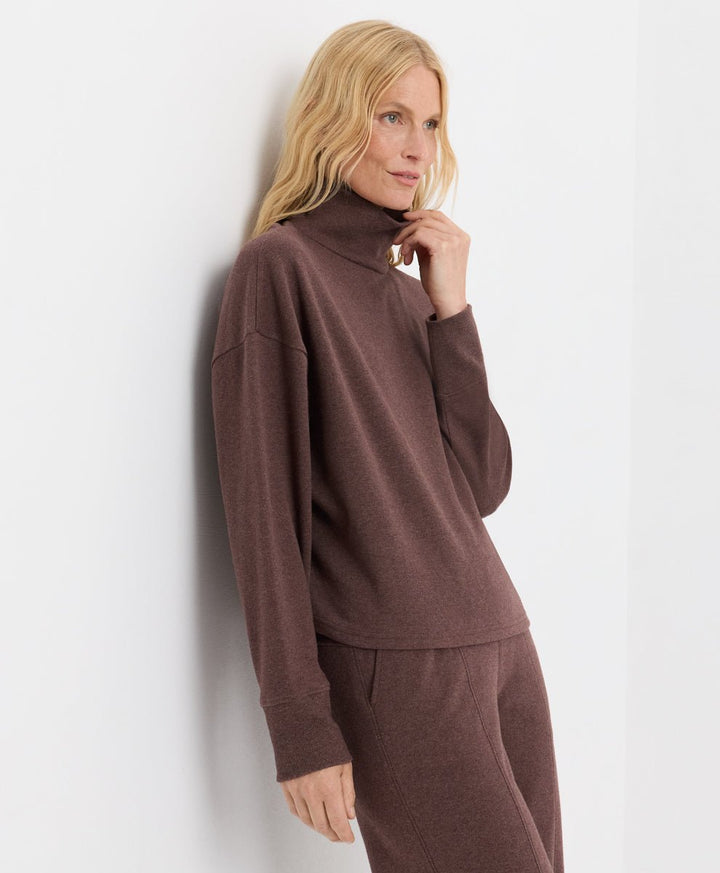 A blonde woman wearing a matching chestnut heather set with a funnel neck long-sleeve top and pants, leaning against a white wall.