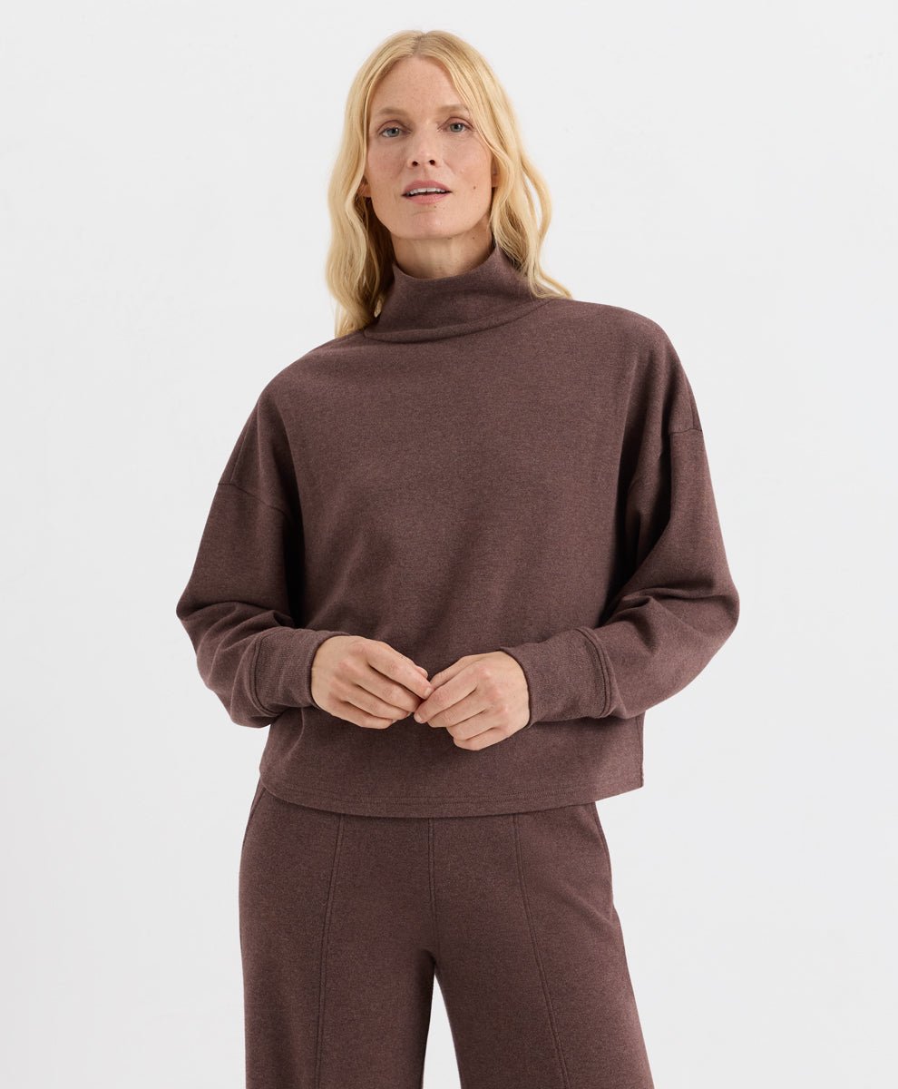 A blonde woman wearing a chestnut heather funnel-neck long-sleeve top and matching brown pants, standing against a light gray studio background.