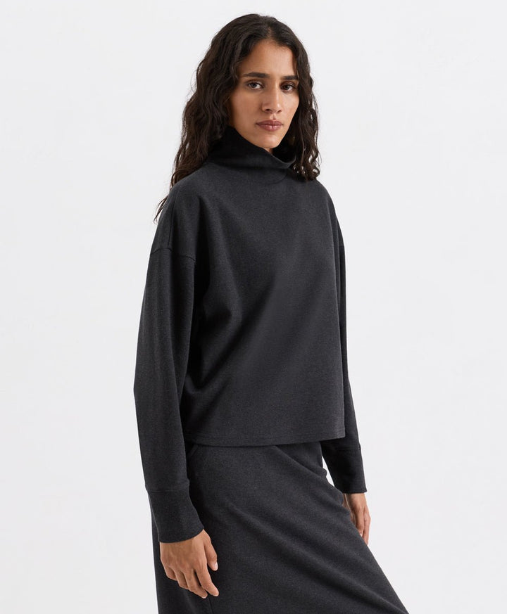 A woman modeling a dark charcoal heather long-sleeve funnel neck top and matching skirt against a plain light gray studio background.