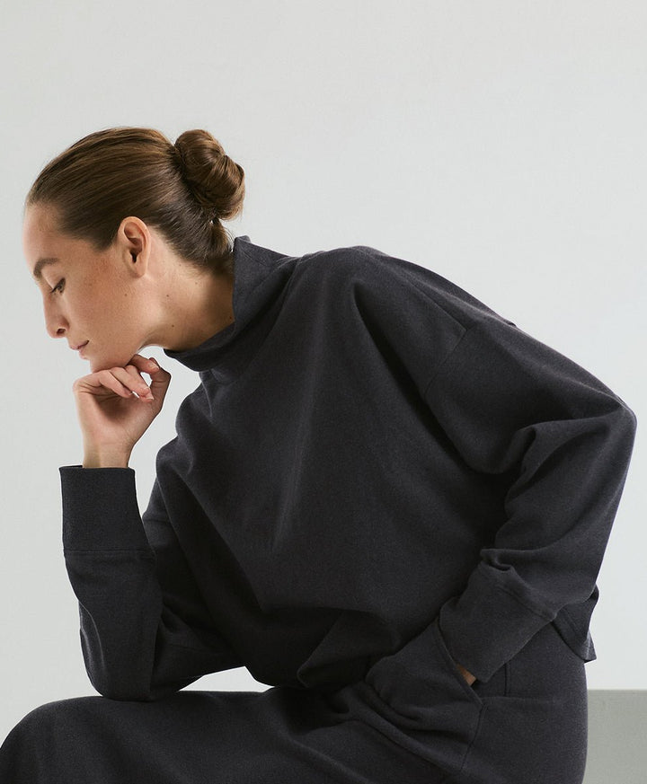 A woman wearing an oversized dark charcoal knit funnel-neck top, posed in a studio setting against a light gray background.
