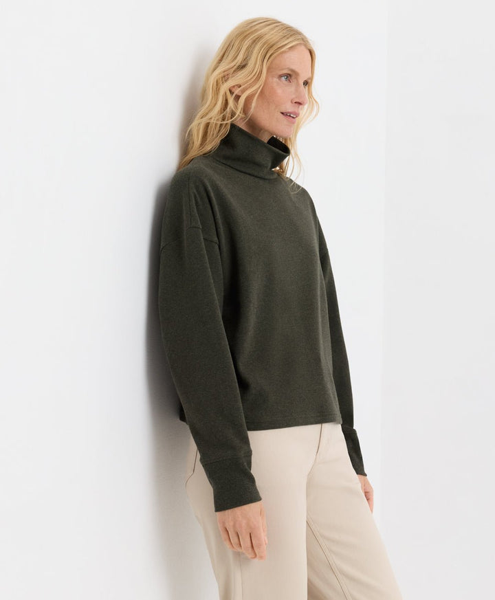 A woman with blonde hair wearing a loden green funnel neck sweater and beige trousers, leaning against a white wall.