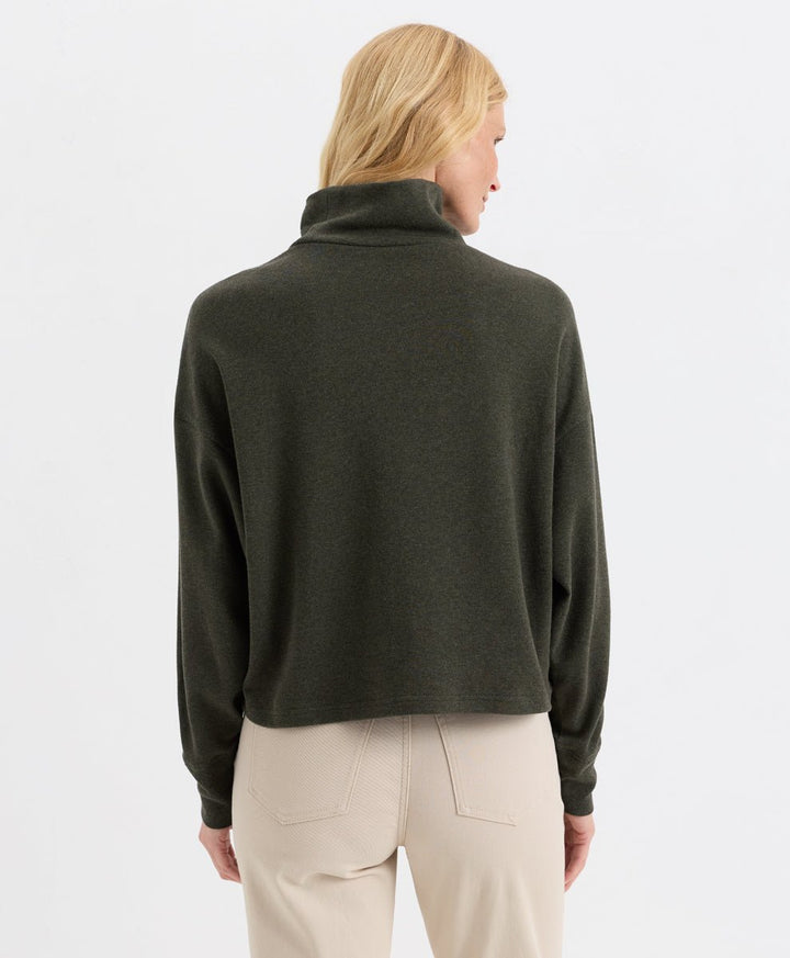 A woman shown from the back wearing a loden green funnel-neck top and light beige trousers, against a plain white studio background.