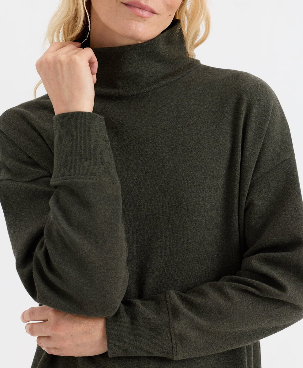 A close-up of a woman wearing a dark olive green ribbed knit turtleneck sweater against a plain white background.