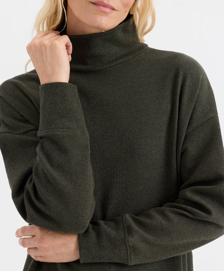 A close-up of a woman wearing a dark olive green ribbed knit turtleneck sweater against a plain white background.