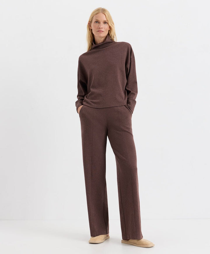 A woman modeling a matching chestnut heather knit lounge set consisting of a long-sleeve mock neck top and wide-leg trousers, paired with beige slip-on shoes against a plain white studio background.