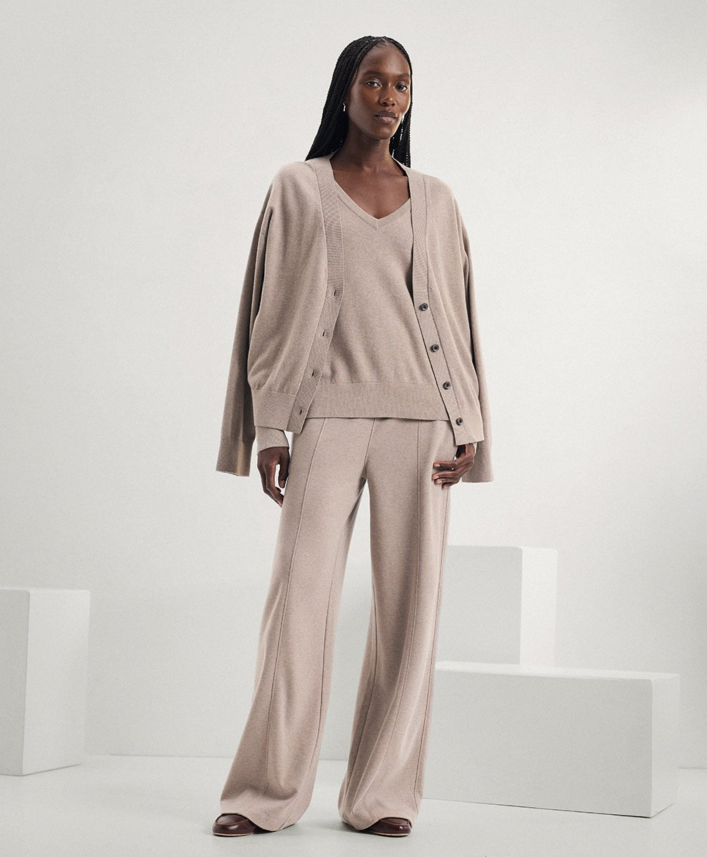 A model stands in a minimalist studio setting wearing a matching beige knit lounge set consisting of a cardigan, V-neck sweater, and wide-leg trousers.
