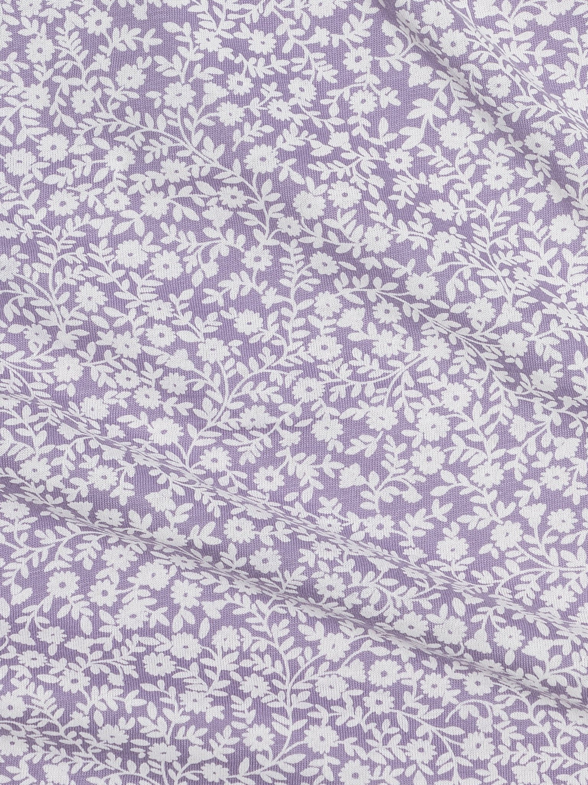 A close-up of a lavender purple fabric with a repeating white floral and leafy vine pattern, shown with soft folds.