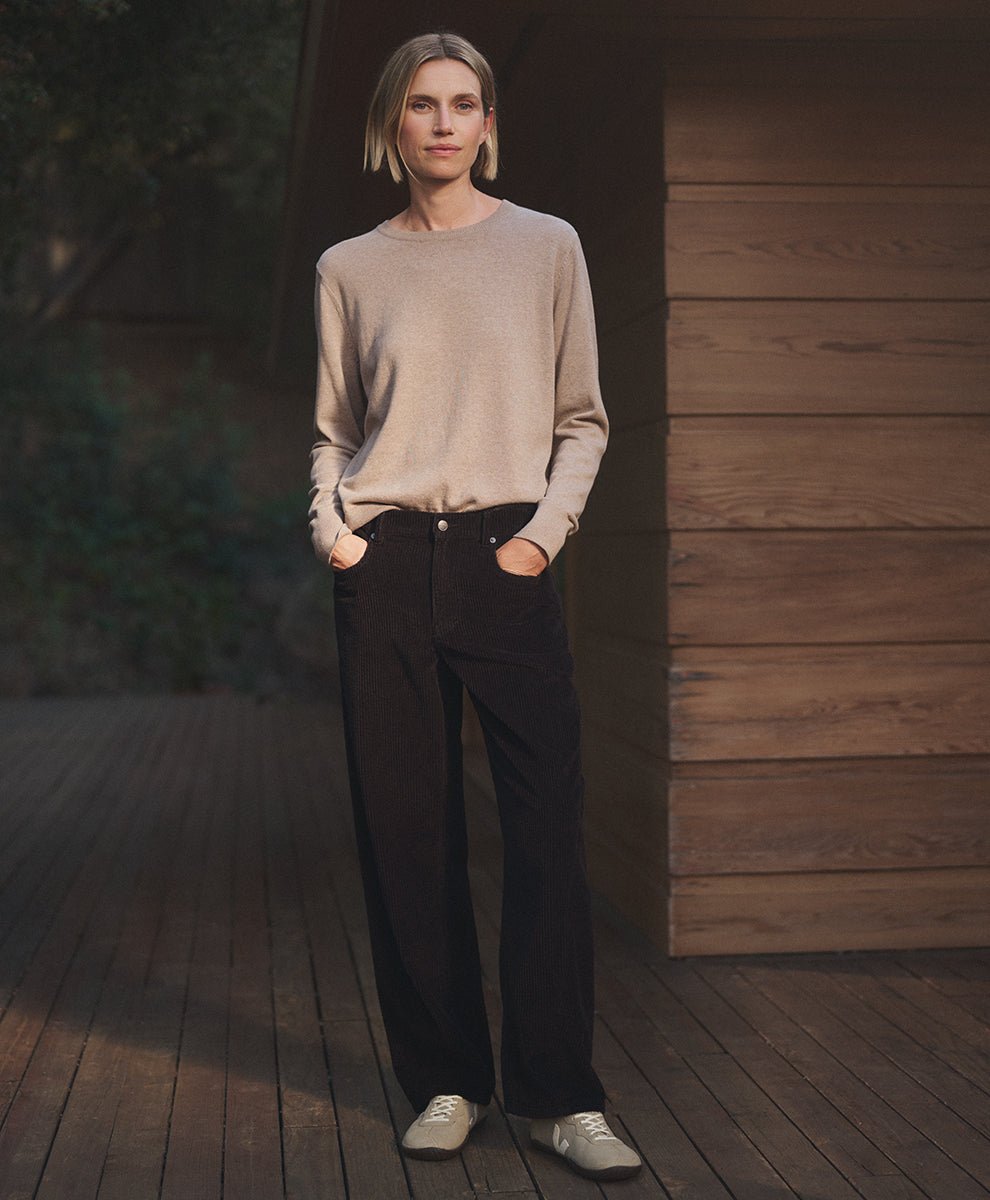 A woman stands on a wooden deck wearing a light beige crewneck sweater, espresso barrel pants, and white sneakers, with a wooden wall and dark background behind her.