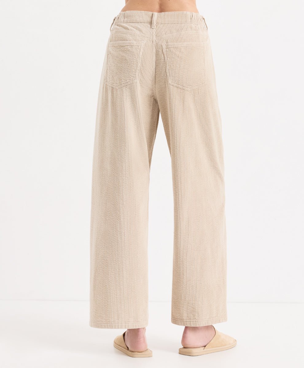Back view of a person wearing high-waisted, corduroy barrel pants in parchment, paired with matching beige slide sandals, against a plain white studio background.