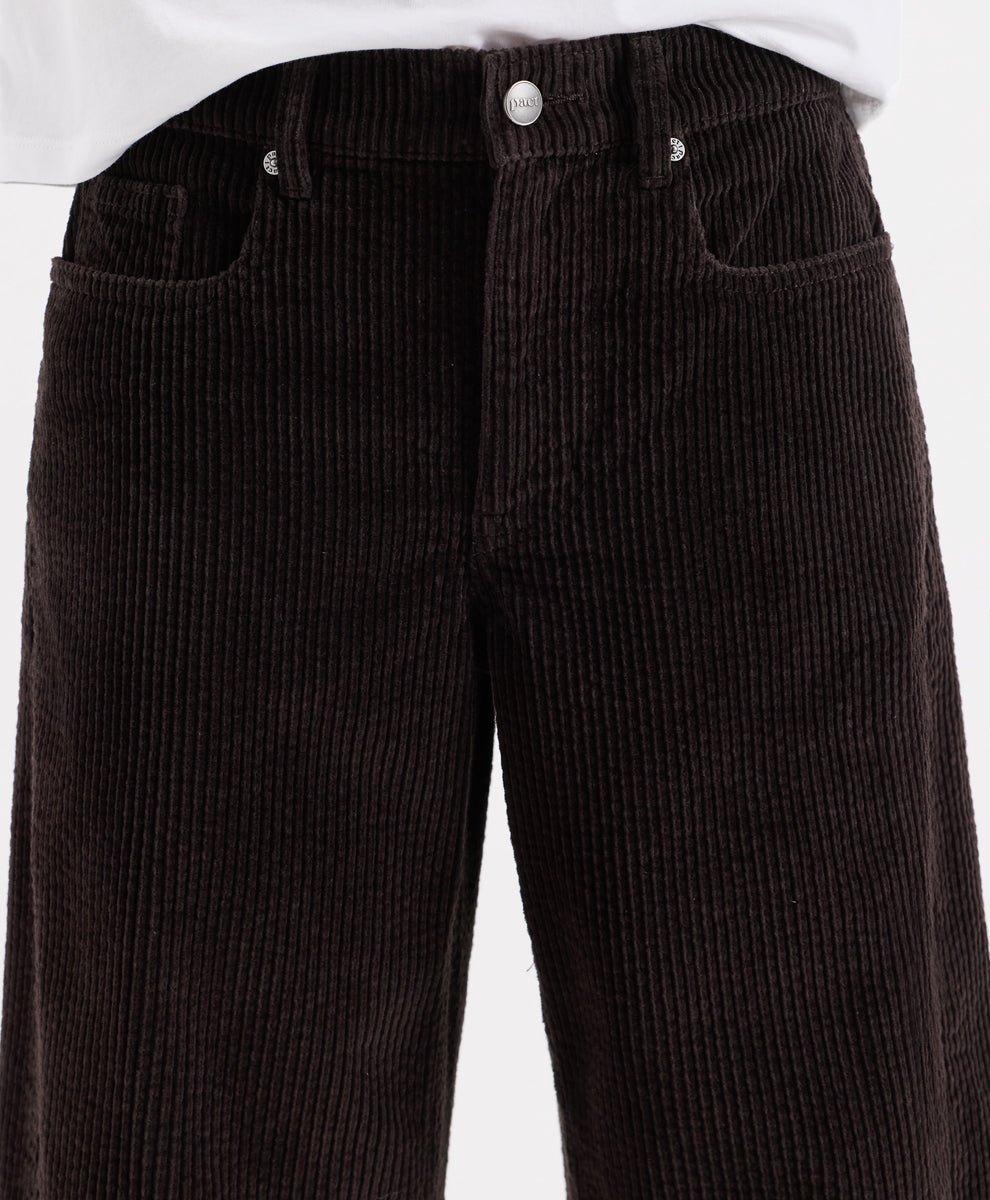 Close-up of espresso corduroy barrel pants with a white shirt hem visible at the top, showing the front waistband, button, belt loops, and side pockets.