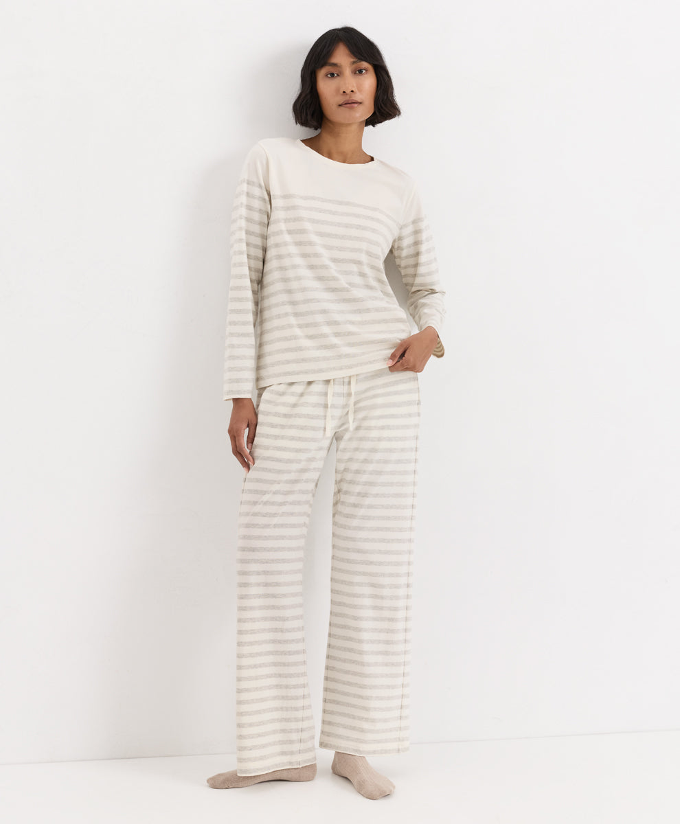 A woman modeling a matching two-piece pajama set consisting of a long-sleeve top and wide-leg pants, both in a cream and light beige horizontal stripe pattern, against a plain white studio background.