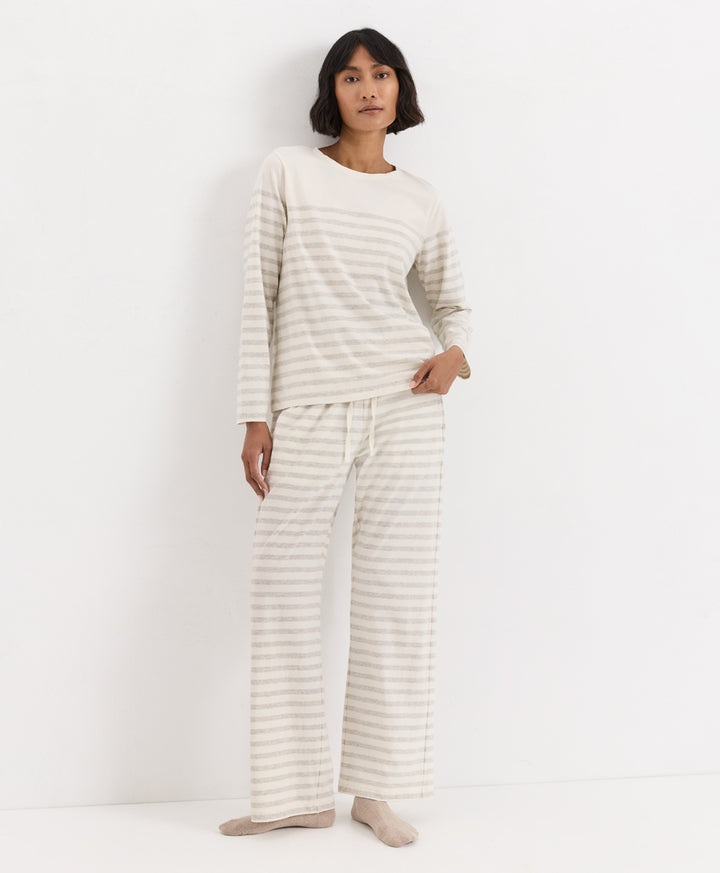 A woman modeling a matching two-piece pajama set consisting of a long-sleeve top and wide-leg pants, both in a cream and light beige horizontal stripe pattern, against a plain white studio background.