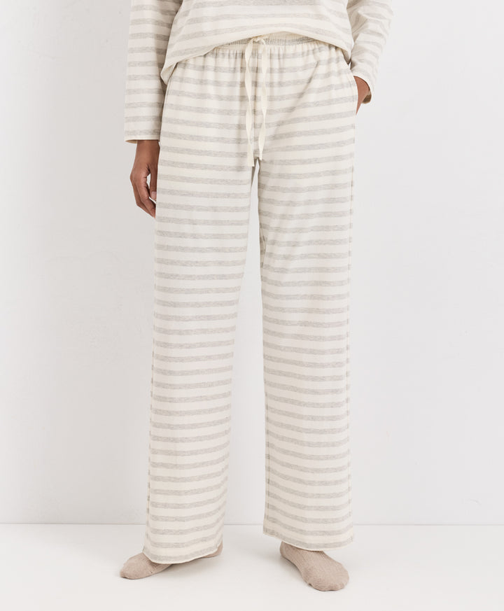 A person wearing loose, wide-leg pajama pants with thin horizontal beige and off-white stripes, paired with a matching long-sleeve top and socks, photographed against a plain light background.