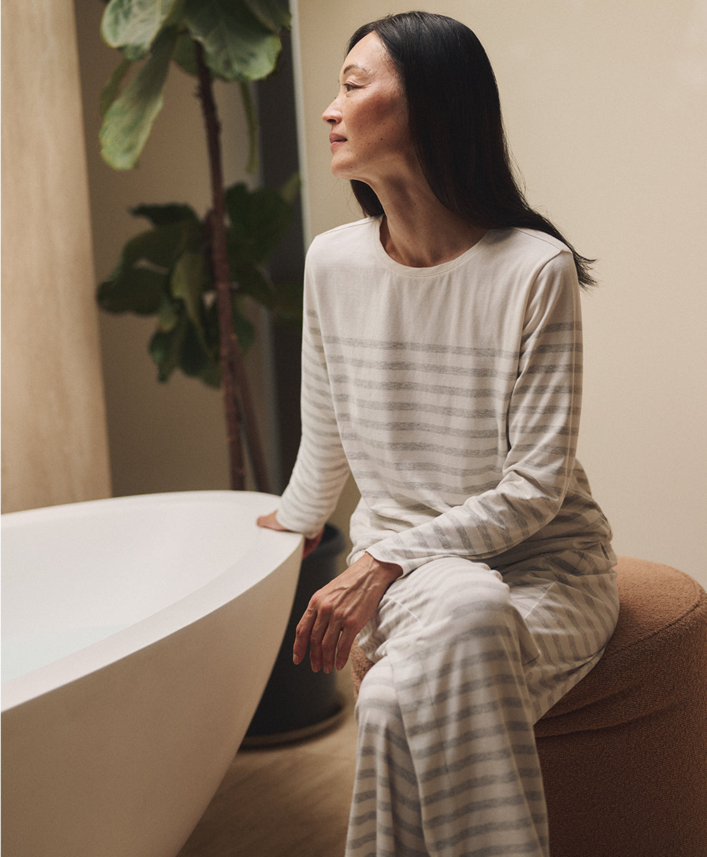 A woman wearing a cream and gray striped long-sleeve pajama set sits on a brown pouf next to a white bathtub in a softly lit bathroom.