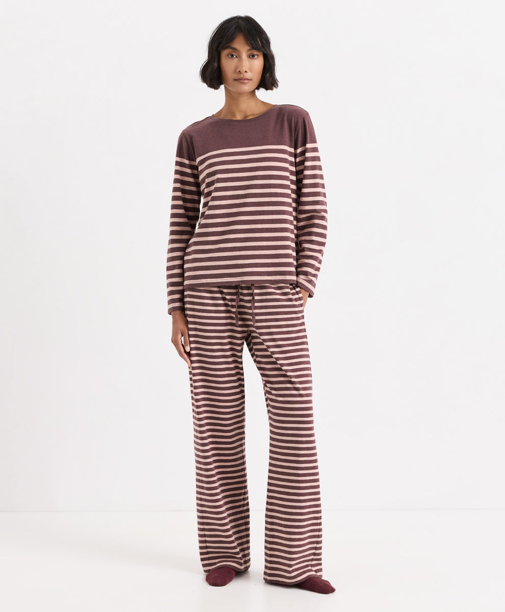 A model stands against a plain white studio background wearing a matching two-piece loungewear set consisting of a long-sleeve top and wide-leg pants, both featuring horizontal stripes in merlot and clay.
